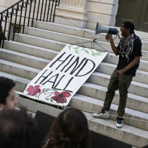 A man with a megaphone on the steps outside of a building with a sign reading Hind Hall